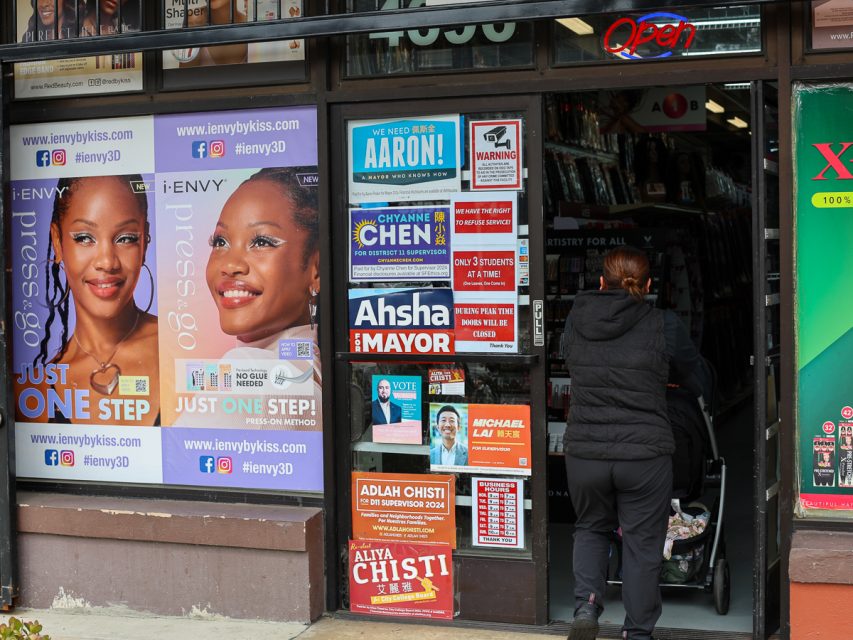 一個商店入口，玻璃窗上有各種選舉海報和廣告。一位穿著黑色外套的人正推著嬰兒車進店。門上方亮著一個霓虹「營業中」標誌。