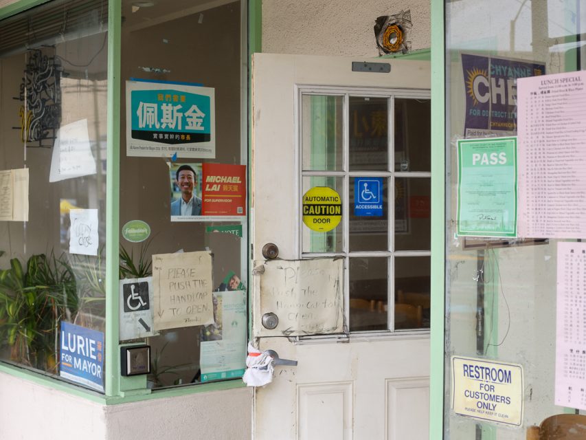 一扇店面門，上面有各種標誌，包括政治海報、無障礙貼紙、洗手間告示，以及一個要求推門把手的標誌。門內可見一株小盆栽。