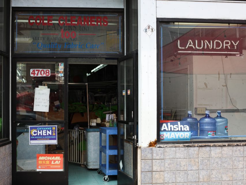 一個店面，上面有「Cole Cleaners」和「洗衣店」的標誌。門上貼有政治競選海報和手寫的「營業時間」便條。店內可見裝水的分水器瓶。