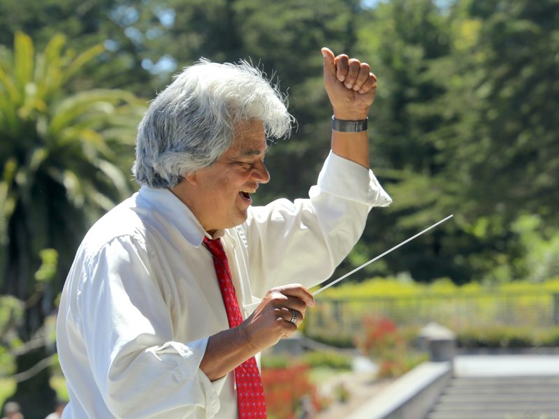 A man in a white shirt and red tie is conducting with a baton, outside on a sunny day with trees in the background.