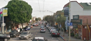 A street view shows parked cars, buildings with storefronts, and a tree-lined roadway extending into the distance under a cloudy sky.