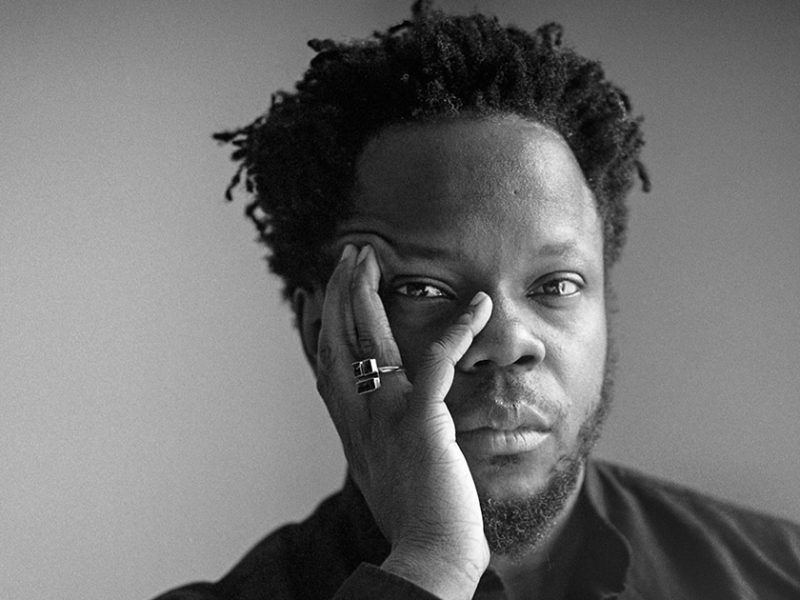Close-up black and white portrait of a person with their hand resting on their face, looking directly at the camera. The individual has short curly hair and is wearing rings on their fingers. This evocative image captures a raw, introspective moment akin to the soulful depth found in Ambrose Akinmusire's music.