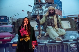 A woman speaks into a microphone in front of an image of a man holding a rifle while sitting on a military vehicle.