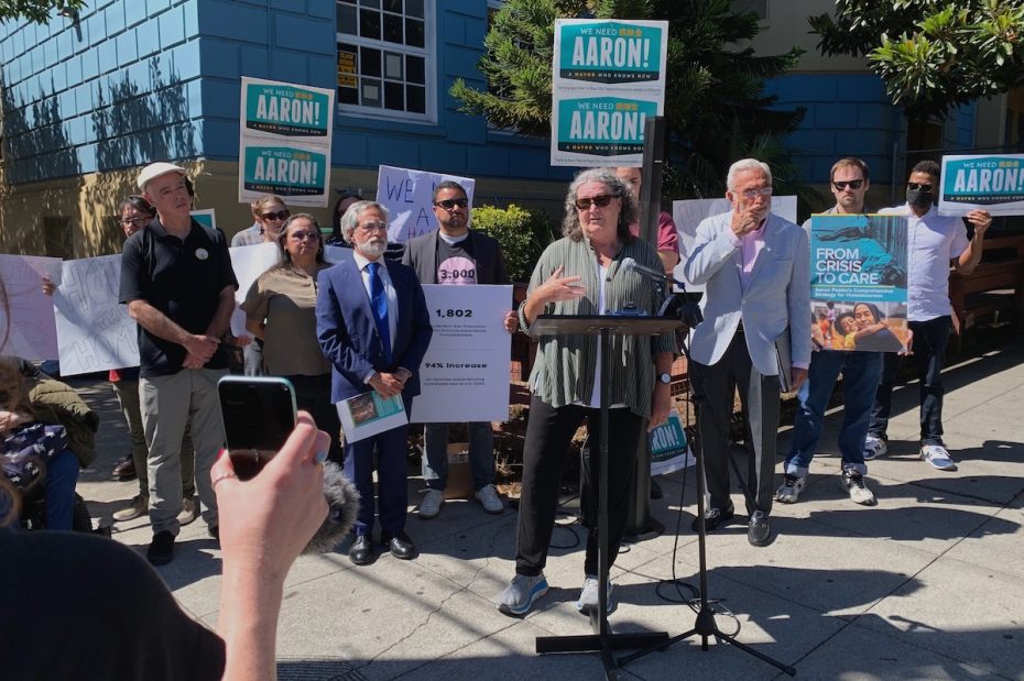 A group of people stand outdoors holding signs, some of which read "We Need This Change" and "From Crisis to Care." A person speaks at a podium. A person in the foreground is holding a smartphone.