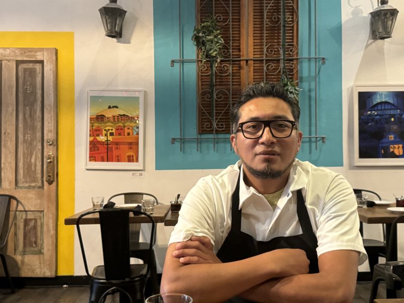 A man with glasses and an apron sits at a table in a brightly colored cafe with artwork on the walls behind him.