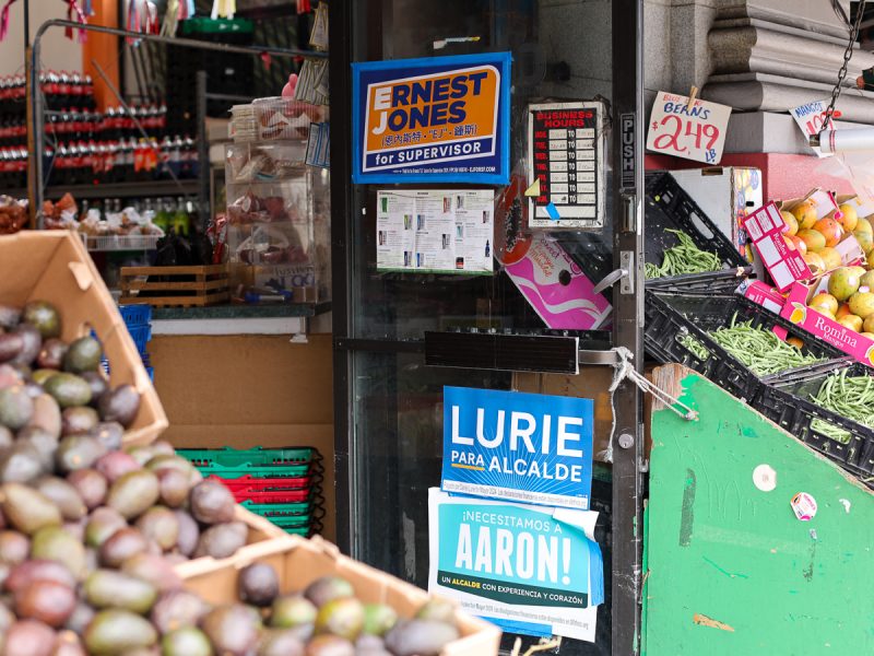 A storefront with political campaign posters on the glass door. Boxes of avocados and beans are displayed outside. Shelves with various items are visible inside the store.