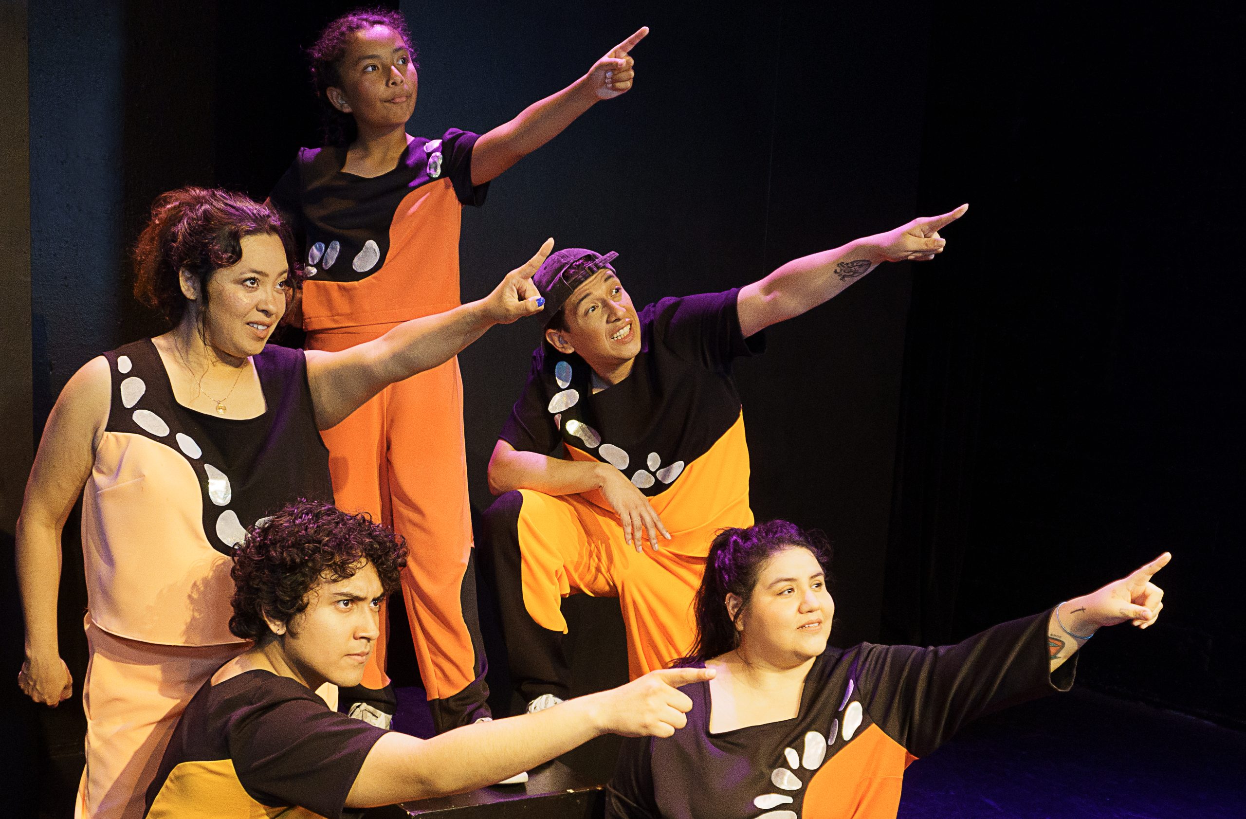 Five people in coordinated orange and black outfits are on a stage, all pointing in the same direction with determined expressions, embodying the spirit of Spanish language theater.