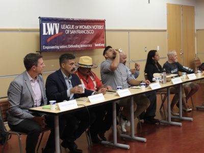 A panel of seven individuals sit at a long table during a meeting. A banner behind them reads "League of Women Voters of San Francisco". One person is drinking from a bottle.