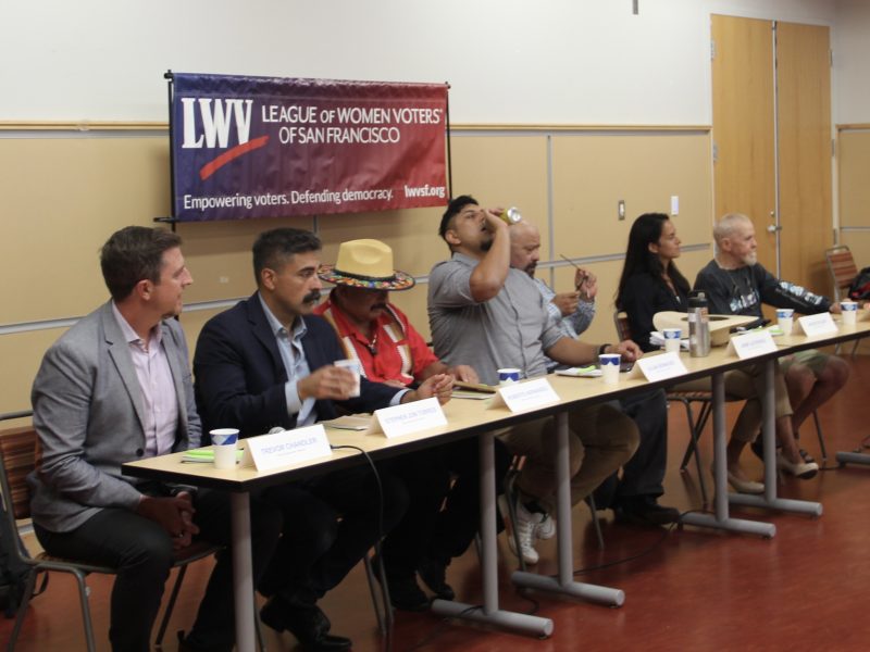 A panel of seven individuals sit at a long table during a meeting. A banner behind them reads "League of Women Voters of San Francisco". One person is drinking from a bottle.