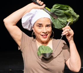 A person in a chef's hat and brown shirt is holding a large leaf and a ladle, positioning them in a playful manner as if wearing them.