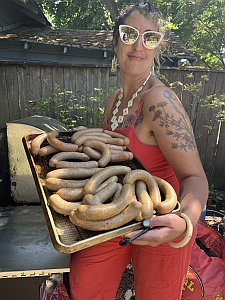 A person in sunglasses and a red dress holds a tray of sausages while standing outdoors.