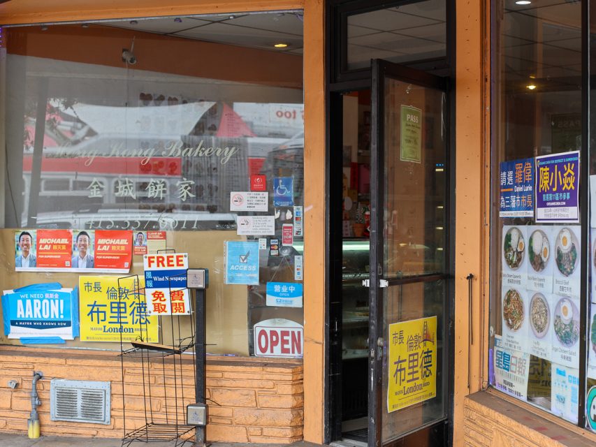 一家麵包店的店面，有黃色的中文字招牌，各種競選海報，以及開著的門。右側窗戶上展示著菜單項目。可見「營業中」的標誌。