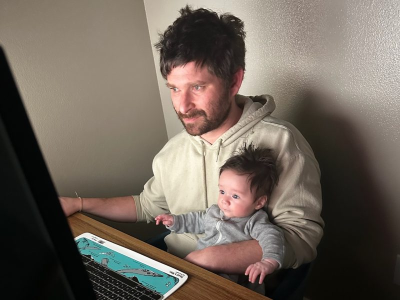A man sits at a desk working on a computer while holding a baby on his lap. Both are looking at the screen.