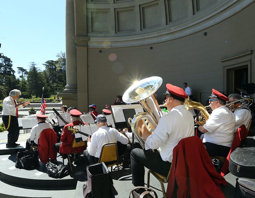 A brass band, wearing red caps and white shirts, performs on an outdoor stage. The conductor stands on the left, leading the musicians. The background features classical architecture and greenery.