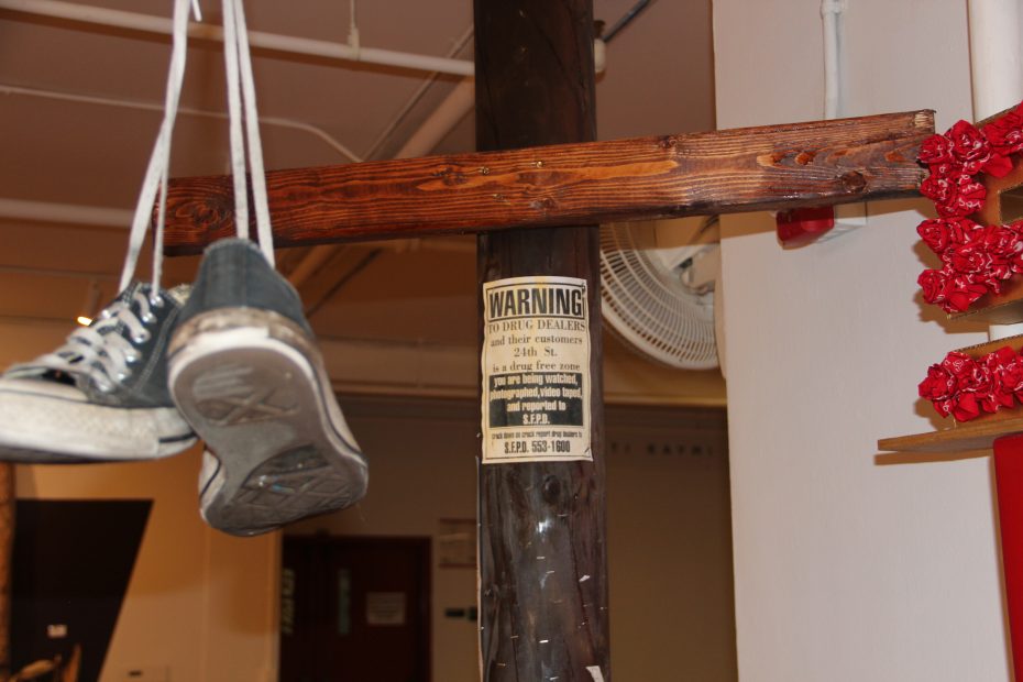 A pair of sneakers hang by their laces from a wooden beam, next to a warning sign on a pole.