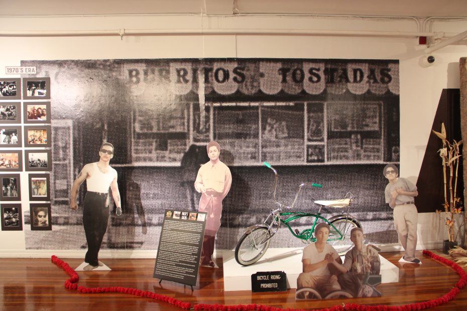Exhibit with life-size cutouts of people, a vintage bicycle, photos, and a sign reading "Burritos Tostadas" in a gallery setting with polished wooden floors.