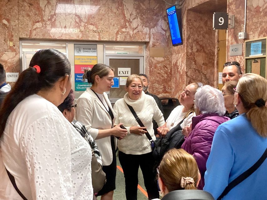 A diverse group of people gathers in a hallway, some engaged in conversation. Signs with directional information are visible on the wall behind them.