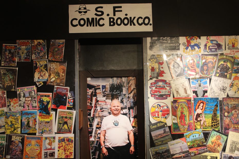 A person stands in the doorway of a comic book store, surrounded by displayed comic books, with a sign above reading "S.F. Comic Book Co.