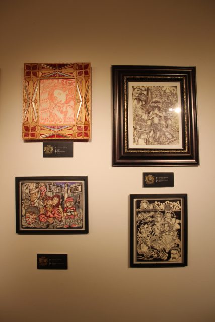 A wall displaying four framed artworks with labels beneath each. The frames vary in size and design, showcasing different artistic styles. The wall lighting highlights the details of each piece.