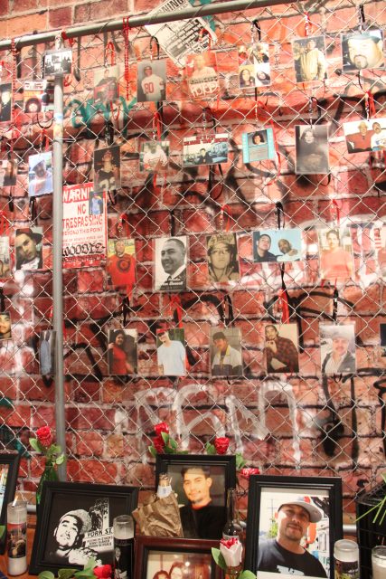 A memorial wall covered in photos and mementos of various people hangs on a brick wall, surrounded by framed pictures, candles, and flower offerings.