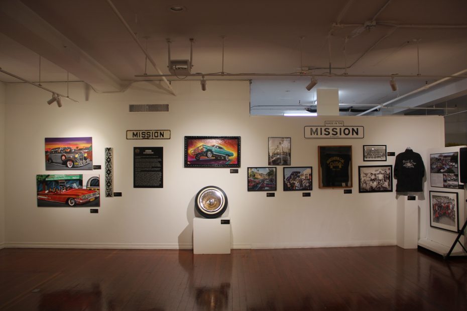 A gallery wall displaying various artworks, photographs, and memorabilia. An informational plaque and a displayed car wheel are also part of the exhibit. The overall theme appears to be automotive.