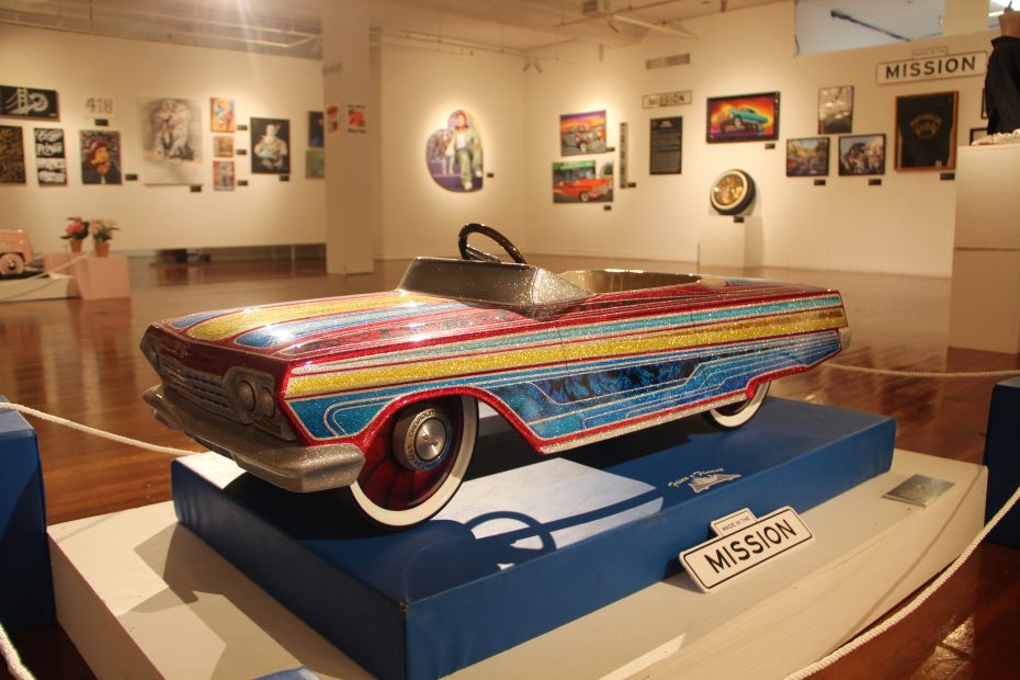 A gallery features various artworks and a colorful, miniature car model displayed on a blue platform labeled "MISSION." The walls are adorned with diverse paintings and art pieces.