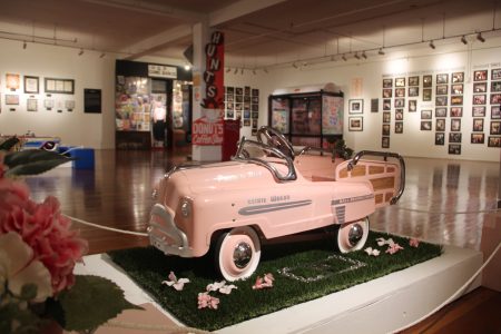 A vintage pink pedal car with "Esther Wagon" written on the side is displayed on grass in a spacious museum gallery filled with various framed pictures and exhibits.
