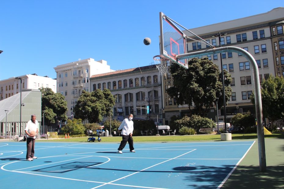 Two people play basketball on an outdoor court surrounded by buildings and trees, with one person shooting the ball towards the hoop.