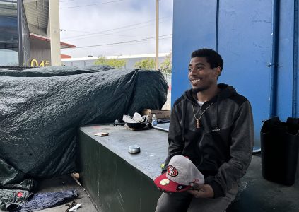 A man wearing a dark hoodie smiles while sitting under a blue overhang. Various items, including hats and bowls, are placed on a green platform next to him. A large tarpaulin covers items in the background.