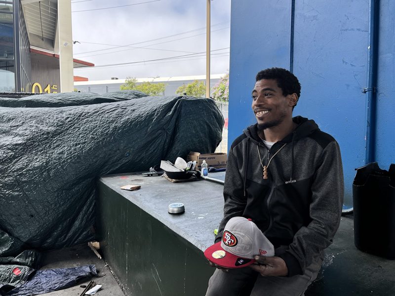 A man wearing a dark hoodie smiles while sitting under a blue overhang. Various items, including hats and bowls, are placed on a green platform next to him. A large tarpaulin covers items in the background.