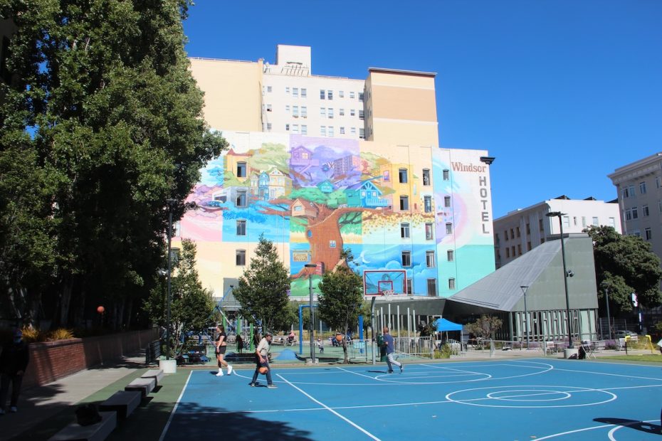 Outdoor basketball court with people playing, overlooked by a building featuring a colorful mural with trees and houses. The building has a sign that reads "Windsor Hotel." Trees surround the court.