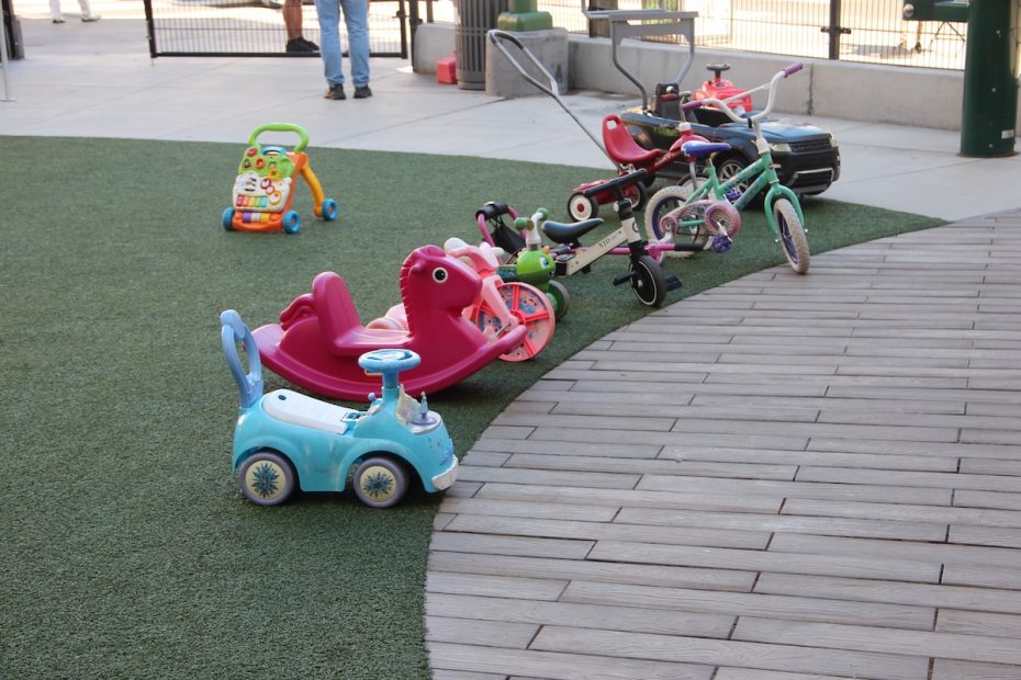 A variety of children's ride-on toys and bikes are scattered on a playground surface, including a blue car and a pink rocking toy.