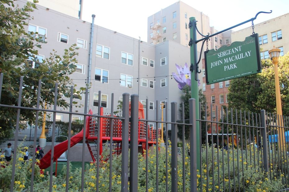 A playground with a red slide is fenced within Sergeant John Macaulay Park, situated next to several apartment buildings. A park sign is visible in the foreground.