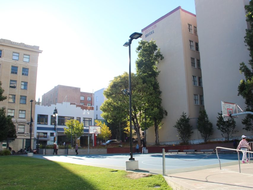 An outdoor basketball court set amidst urban buildings, with a lamppost in the center and people scattered around the area. Some greenery and trees are visible along the edges.