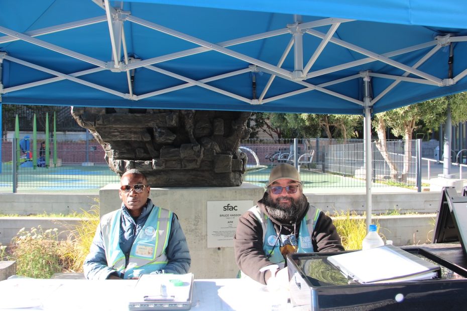 Two people in safety vests sit under a blue canopy tent at an outdoor information booth. They are positioned in front of a playground with a rock structure.