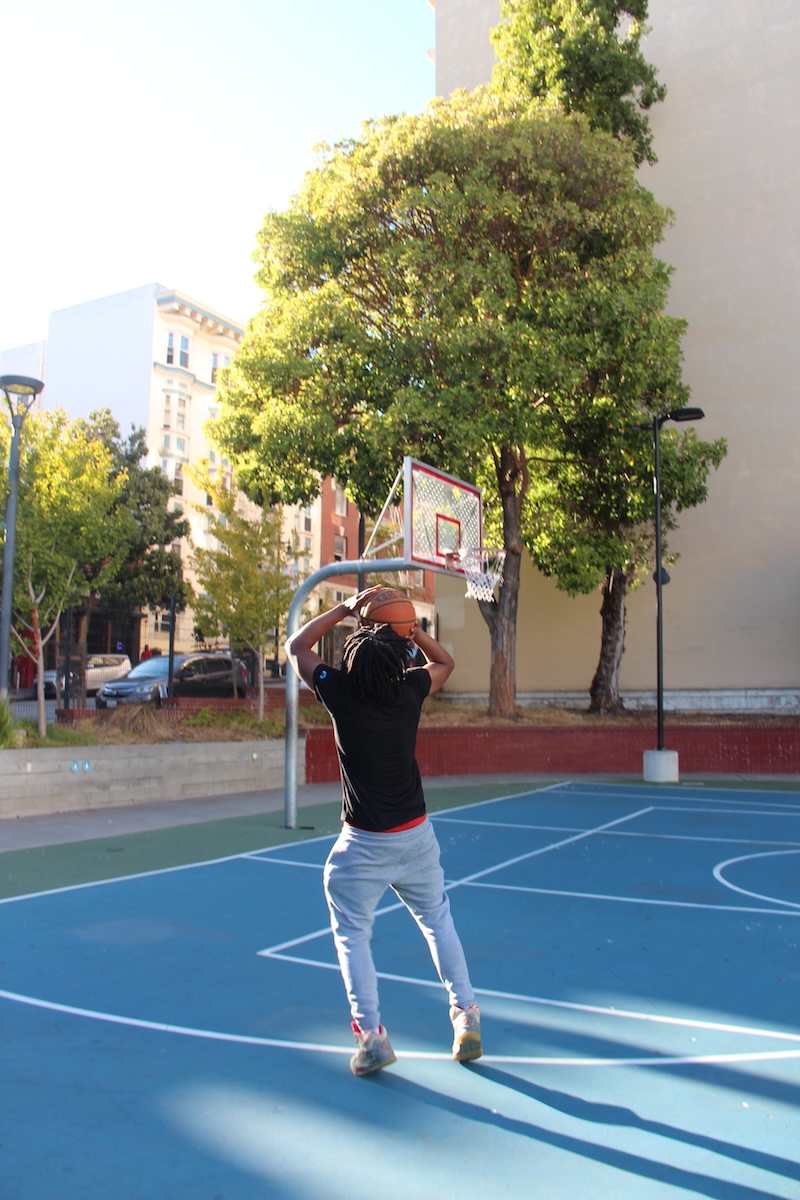 Person in a black shirt and grey pants taking a basketball shot on an outdoor court with blue flooring, surrounded by trees and buildings.