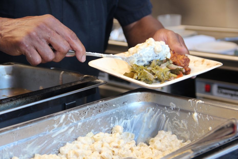 A person serves macaroni and cheese, greens, and a meat dish onto a paper plate at a food service station.