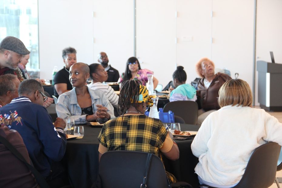 A diverse group of people is seated around tables in a well-lit room, engaged in conversation during a meal.