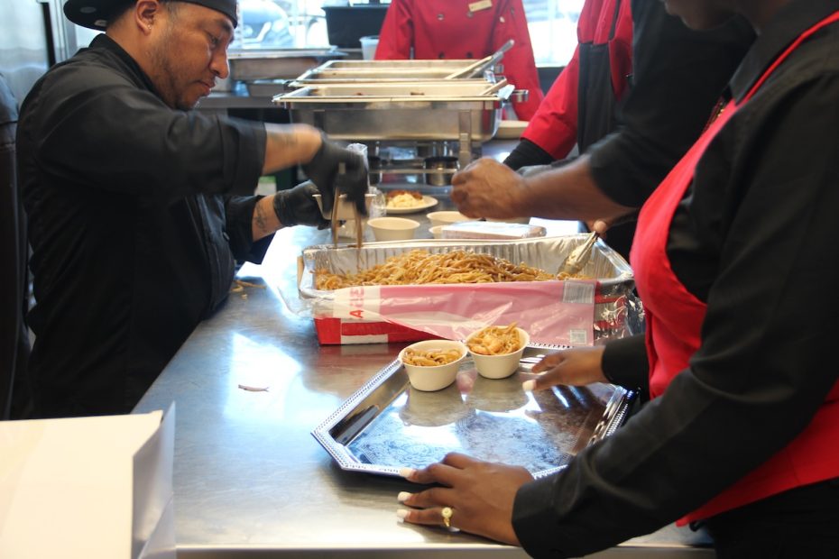 A person in black kitchen attire serves food from a large tray, placing portions into small bowls. Another person in black and red attire holds a tray with bowls of food.