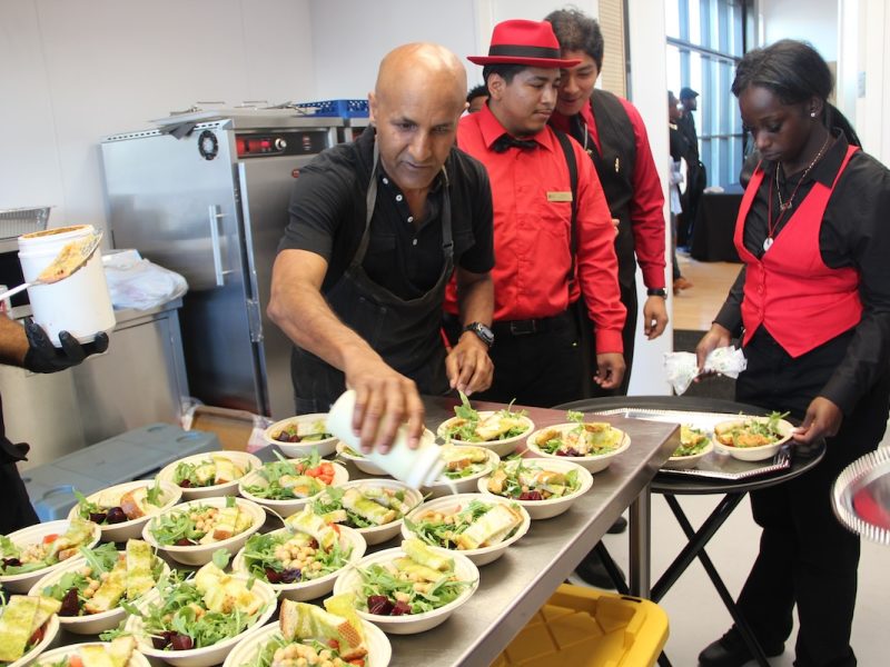 Cooks and servers in black and red uniforms prepare and garnish bowls of salad in a commercial kitchen setting.