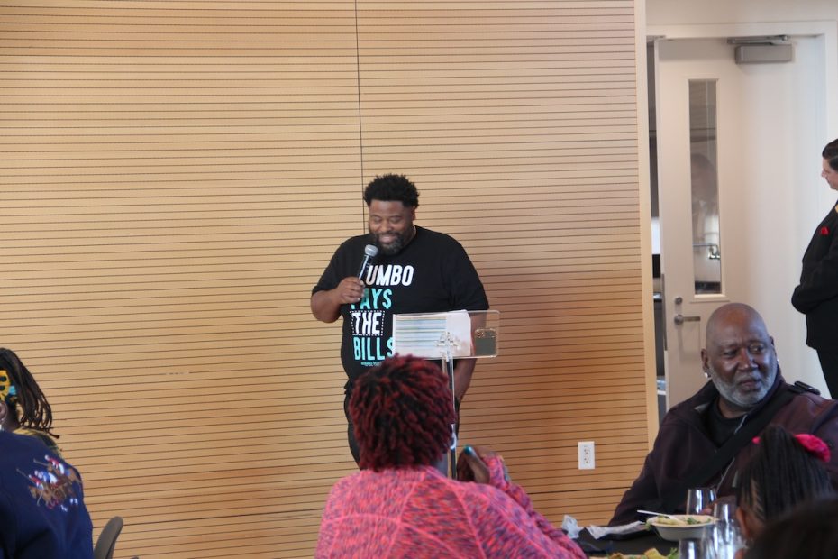 A man is speaking into a microphone at an indoor event. He is wearing a black T-shirt with text on it. Individuals sit at tables, listening attentively, including an older man in the foreground.