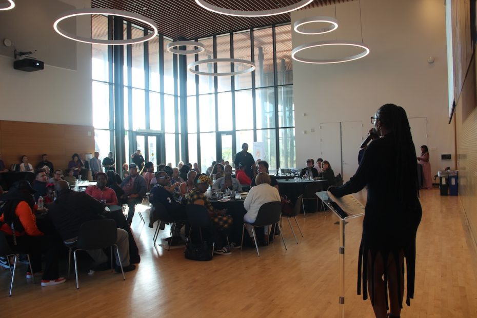 A person speaks at a podium in a modern hall with large windows and circular ceiling lights while a seated audience listens attentively.