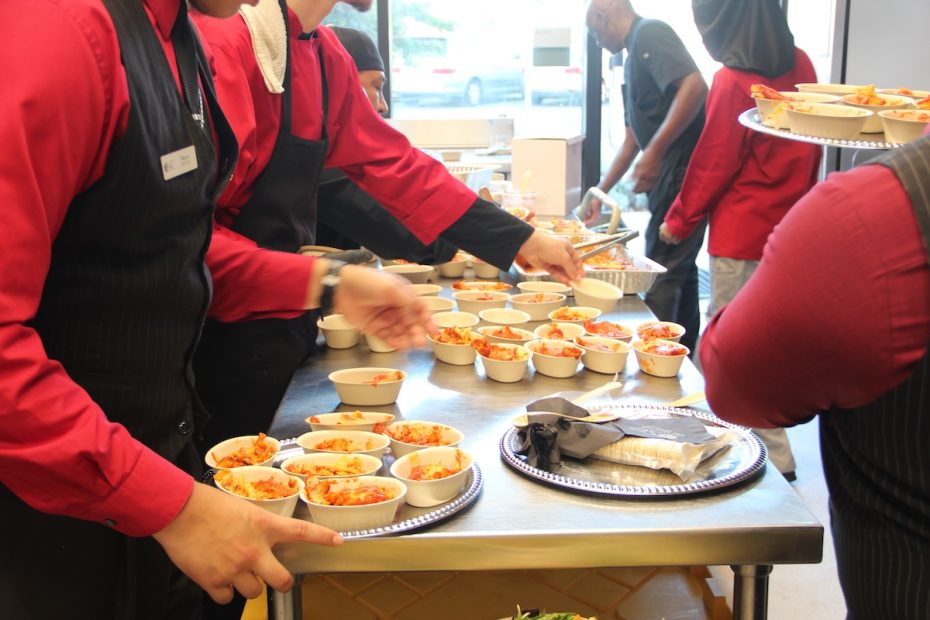 Catering staff in red shirts and black vests are placing bowls of food on a table while a person in the background carries more bowls.