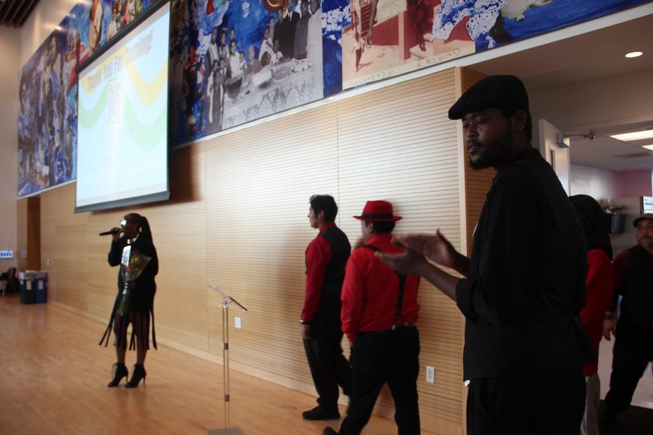A person speaks while four individuals in red and black attire stand nearby, with a mural and large screen in the background.
