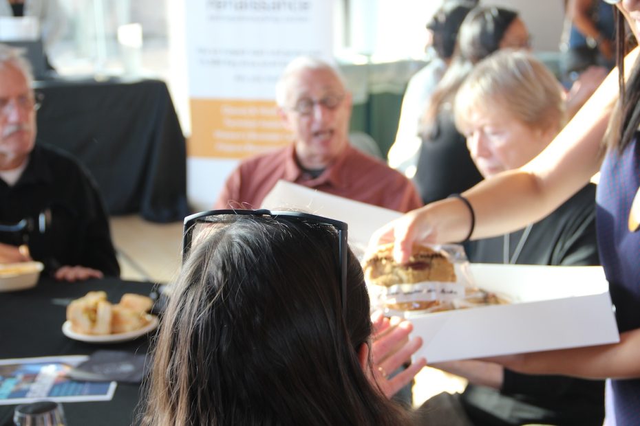A group of people is seated at a table with food. One person is holding an open box with pastries, offering them to the others. Some individuals are focused on the pastries, while others are conversing.