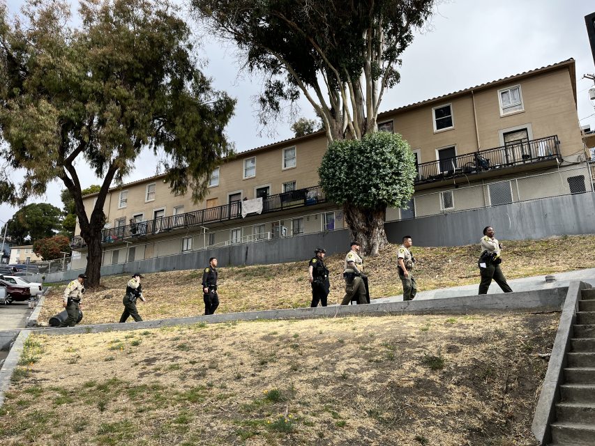 A group of uniformed individuals walk in a line along a grassy slope in front of a row of residential buildings.