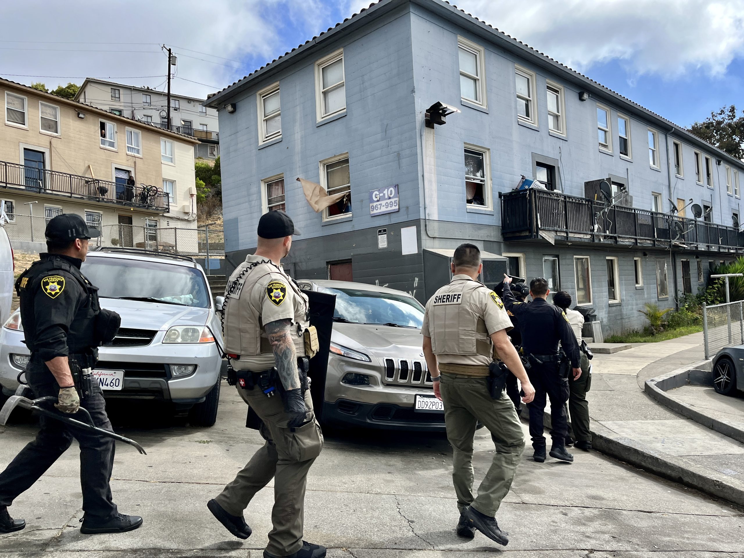 Several sheriff officers in uniform approach a blue residential building with parked vehicles nearby. Some officers hold tactical gear.