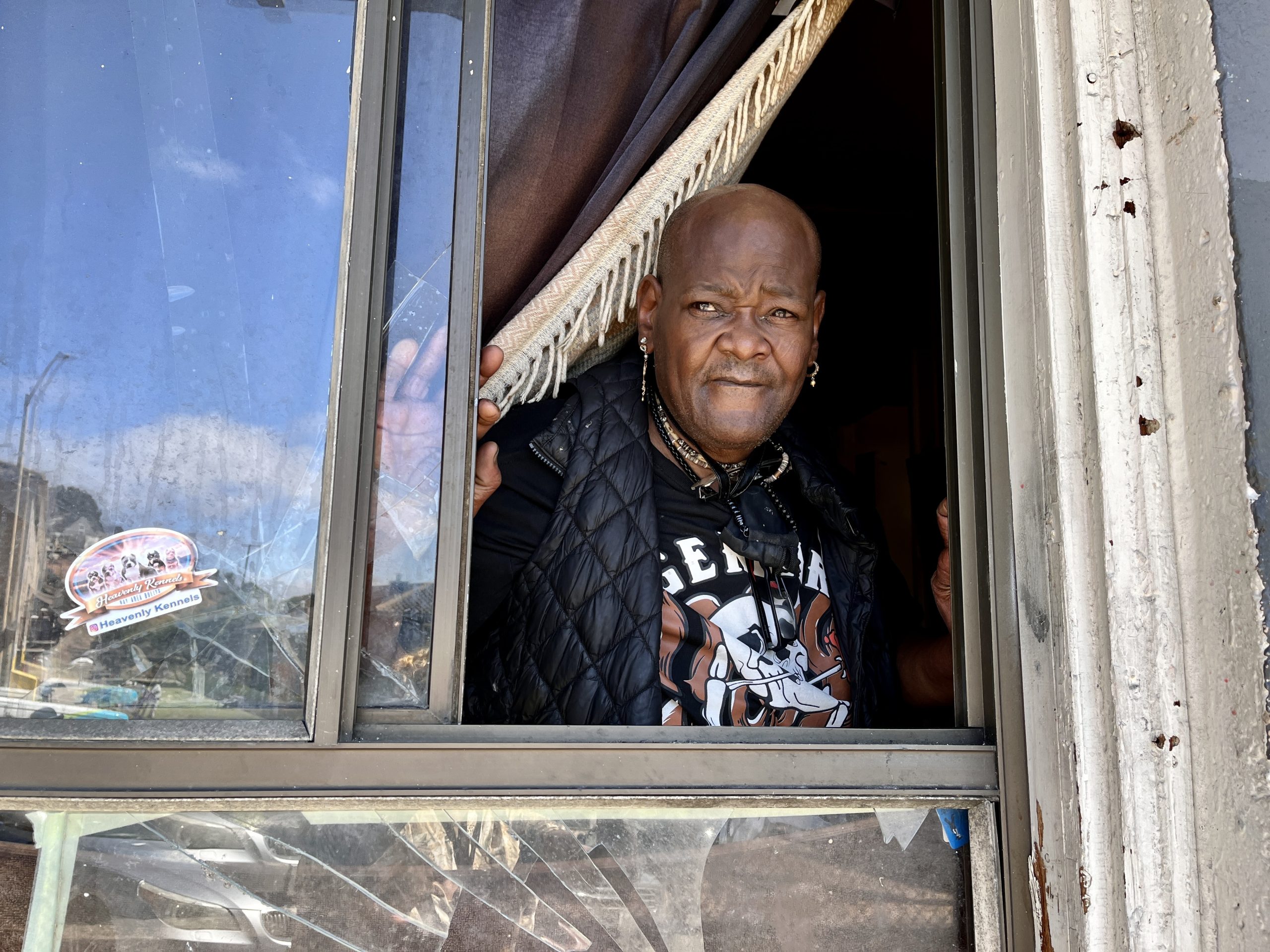 A person looks out from an open window, partially covered by a curtain, with a neutral expression. The window frame appears aged and weathered.