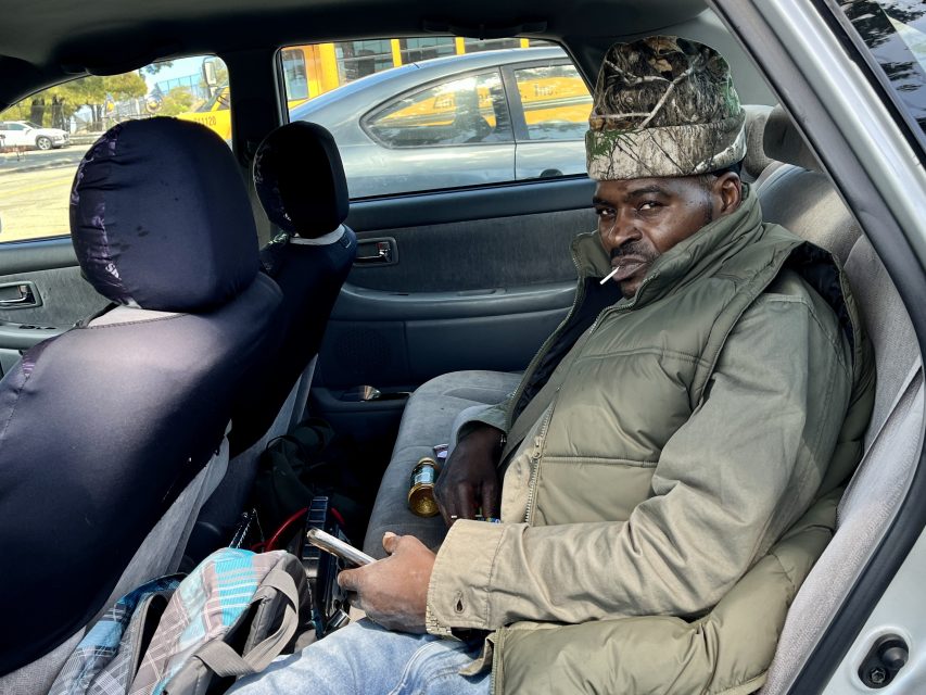 A man wearing a camouflage hat and green jacket reclines in the backseat of a parked car, holding a smartphone.
