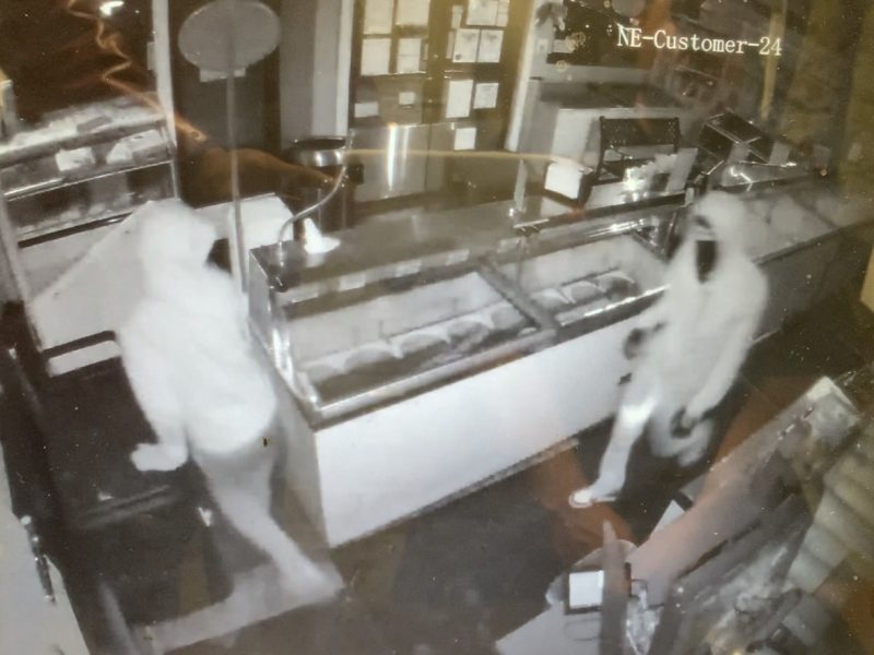 Surveillance footage of two individuals in hooded attire inside a dark, seemingly closed store. One person is near a counter with glass display cases, the other is to the right, both partially obscured.
